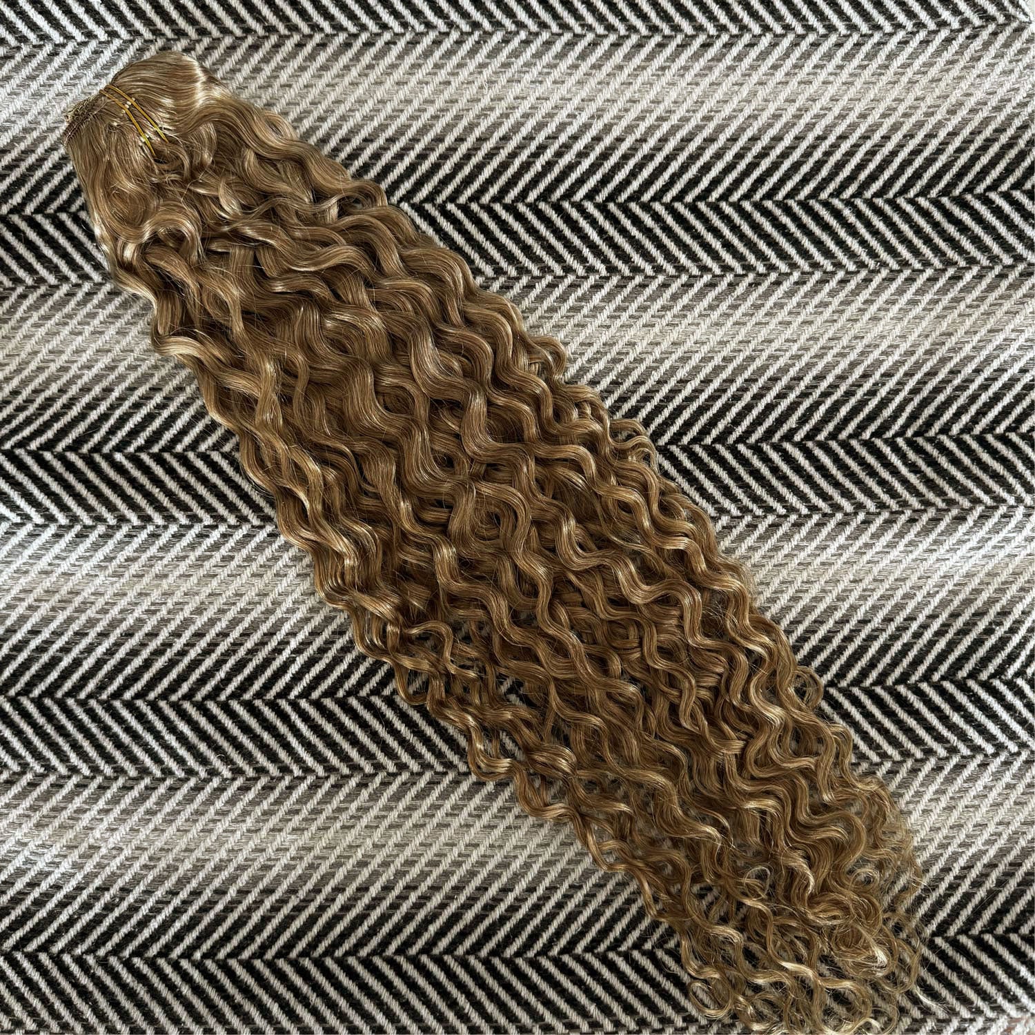 Curly hair extension on a textured fabric background, shade is Dirty Blonde