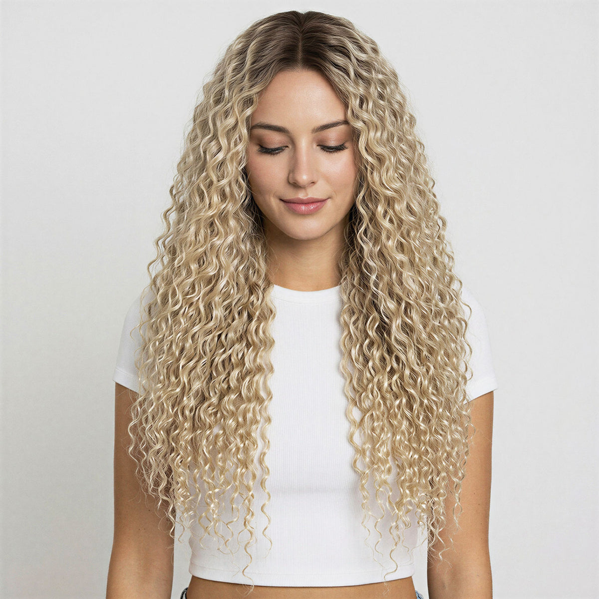 Woman with long, wavy blonde curly hair extensions wearing a white top against a plain background