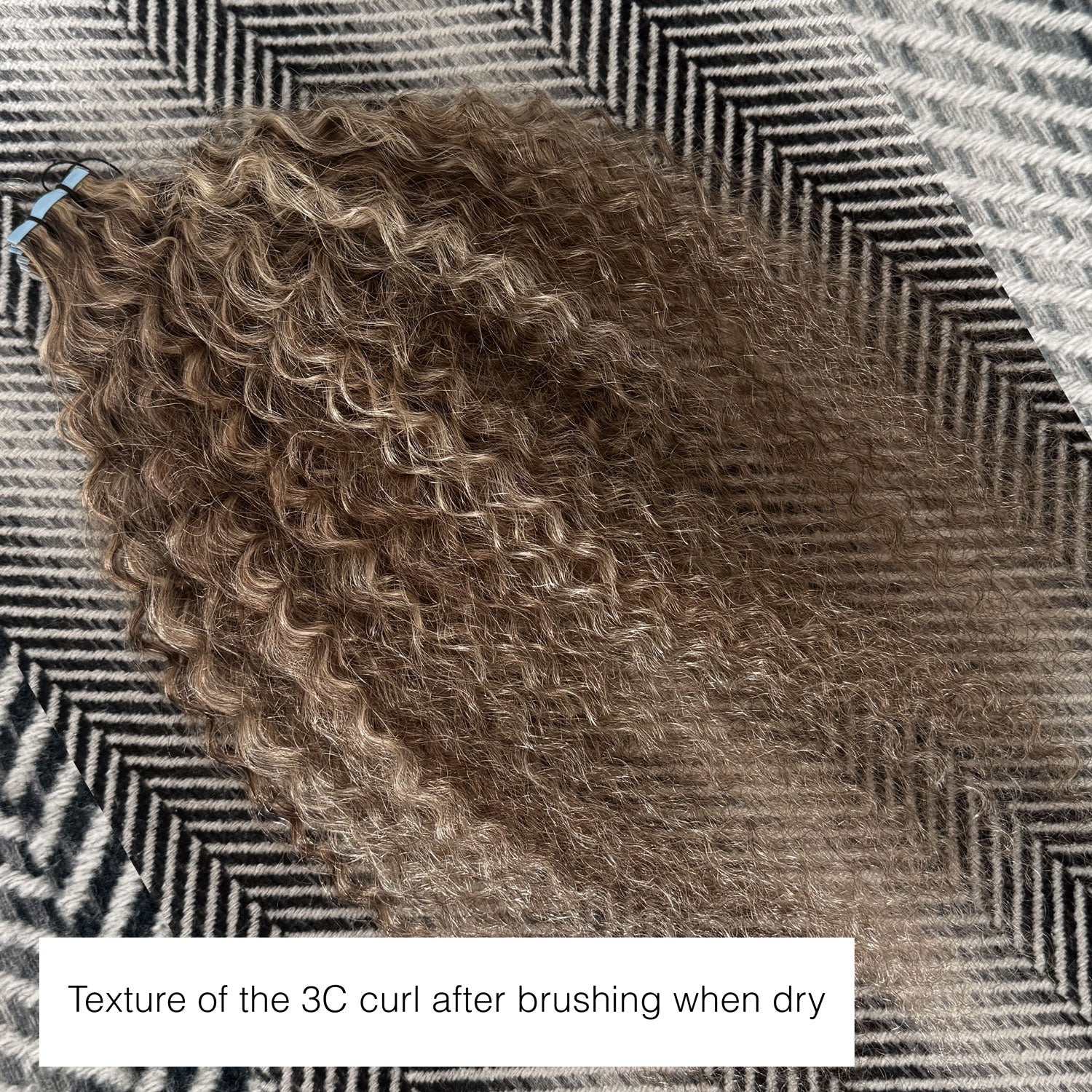 A bundle of 3C curly hair extensions with a brown and blonde ombré color, displayed on a black and white herringbone patterned fabric background. A text overlay at the bottom reads 'Texture of the 3C curl after brushing when dry'.