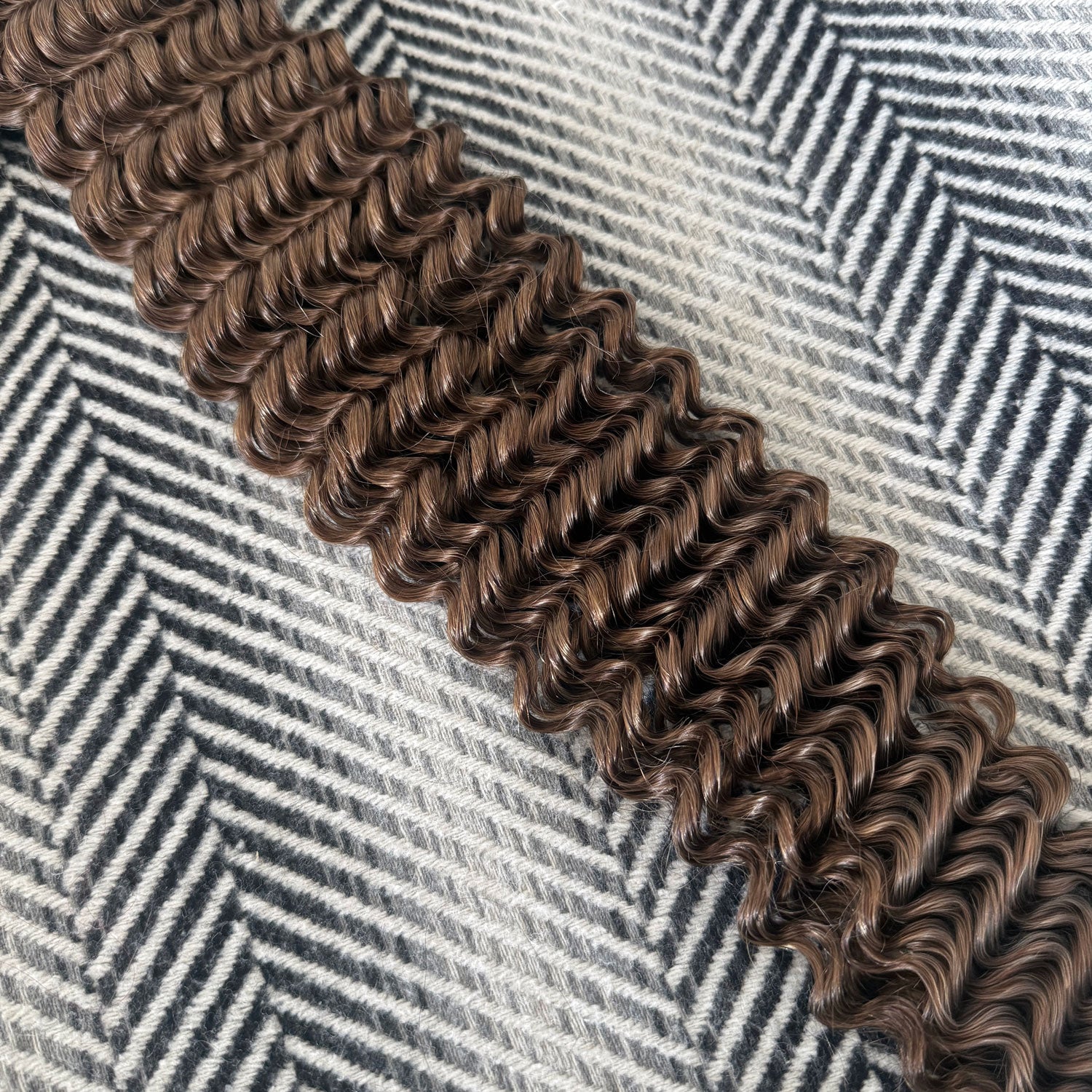 A close-up of a medium brown curly hair extension or wig section laid diagonally across a black and white herringbone patterned fabric background.