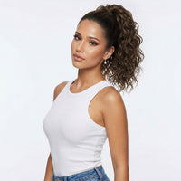 Woman wearing a white tank top and blue jeans with a curly ponytail lagainst a plain background