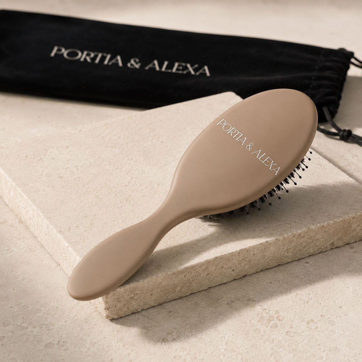 A beige oval hairbrush with black bristles rests on a light stone slab, with a black drawstring pouch printed with the PORTIA & ALEXA brand name in the background.