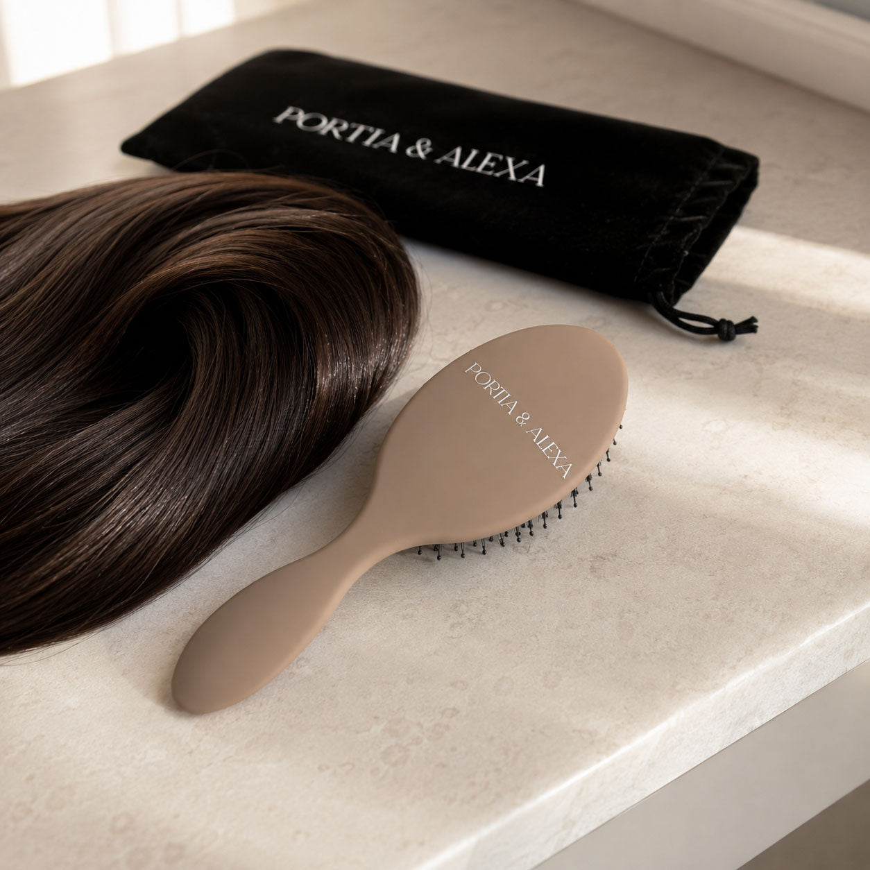 A dark brown hairbrush with a beige handle and black bristles, shown next to a matching black velvet drawstring pouch and a bundle of dark brown hair on a light marble surface. The brand name 'PORTIA & ALEXA' is printed on both the brush and the pouch.