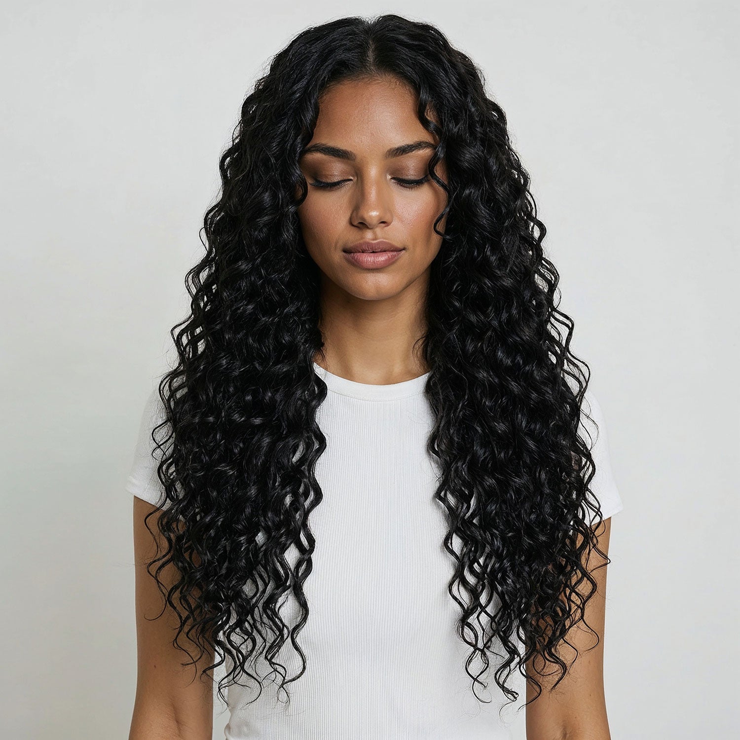 Woman with long, wavy black hair  extensions wearing a white top against a plain background