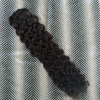 A bundle of dark brown, tightly curled hair extensions or a wig section laid diagonally on a black and white herringbone patterned fabric background.