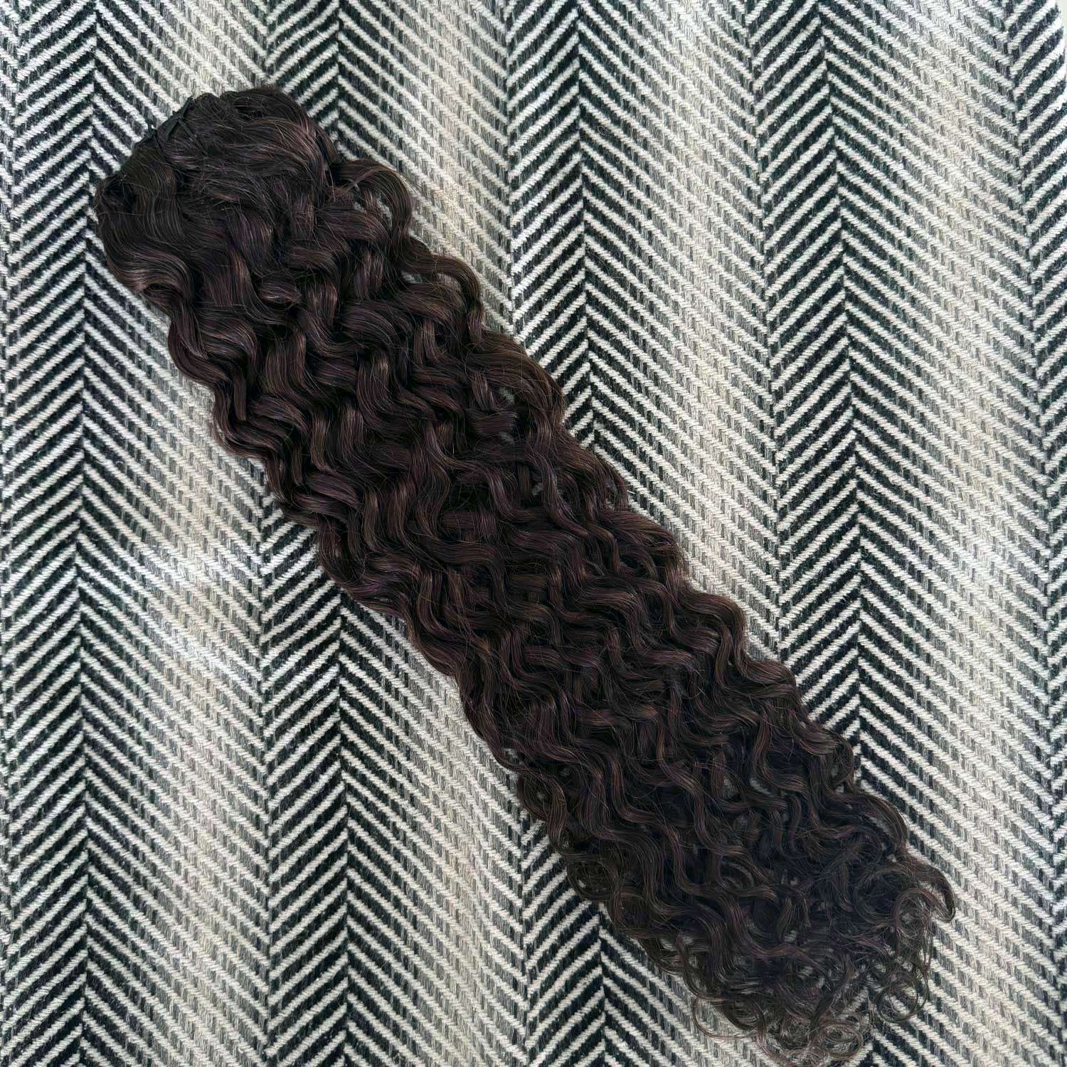 A bundle of dark brown, tightly curled hair extensions or a wig section laid diagonally on a black and white herringbone patterned fabric background.