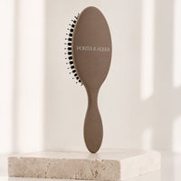 A matte taupe oval hairbrush with black bristles and the brand name 'PORTA & ALEXA' printed on the handle, displayed on a light stone block against a softly lit cream background.