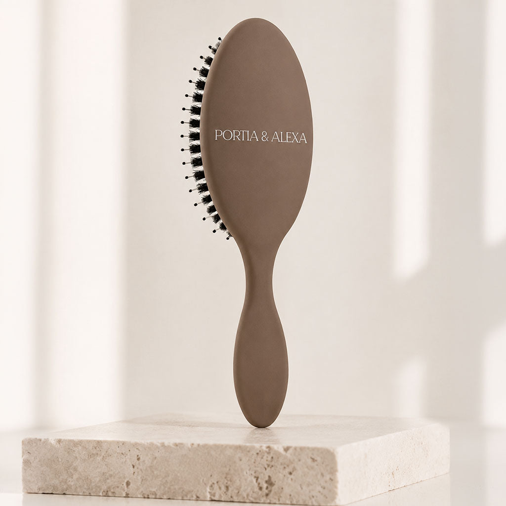A matte taupe oval hairbrush with black bristles and the brand name 'PORTA & ALEXA' printed on the handle, displayed on a light stone block against a softly lit cream background.