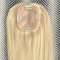 A close-up of a blonde hair extension piece with a light beige adhesive base and four metal clip attachments, laid flat on a gray-and-white patterned fabric background.