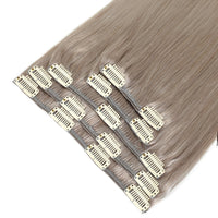 A close-up of straight clip-in hair extensions in a cool ash blonde color, showing multiple hair wefts attached to gray adhesive bands and white metal clip ends, set against a plain white background.