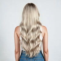 A woman shown from the back with long, wavy hair in a highlighted blonde shade, wearing a white sleeveless top and black bottoms against a plain light gray background.