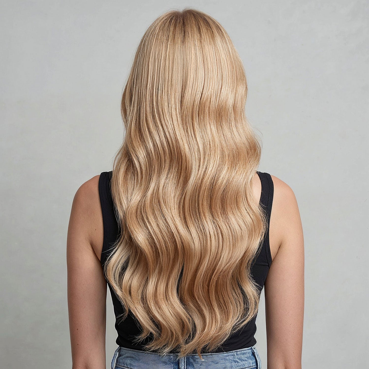 Person with long, wavy honey blonde hair extensions on a gray background