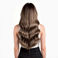 A woman viewed from behind wearing a white sleeveless top and black jeans, showcasing long, wavy hair with a dark brown root and warm blonde highlights.