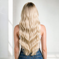 A woman viewed from behind wearing a long hair extensions, wavy blonde wig with dark roots, paired with a white tank top and blue jeans against a bright white background.