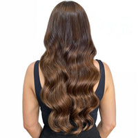 Person with long, wavy brown hair extensions wearing a white shirt and blue jeans on a light gray background