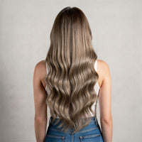 A woman viewed from behind with long, wavy hair, showing a highlighted blonde-to-brown ombre style. She is wearing a white tank top and blue jeans against a plain light gray background.