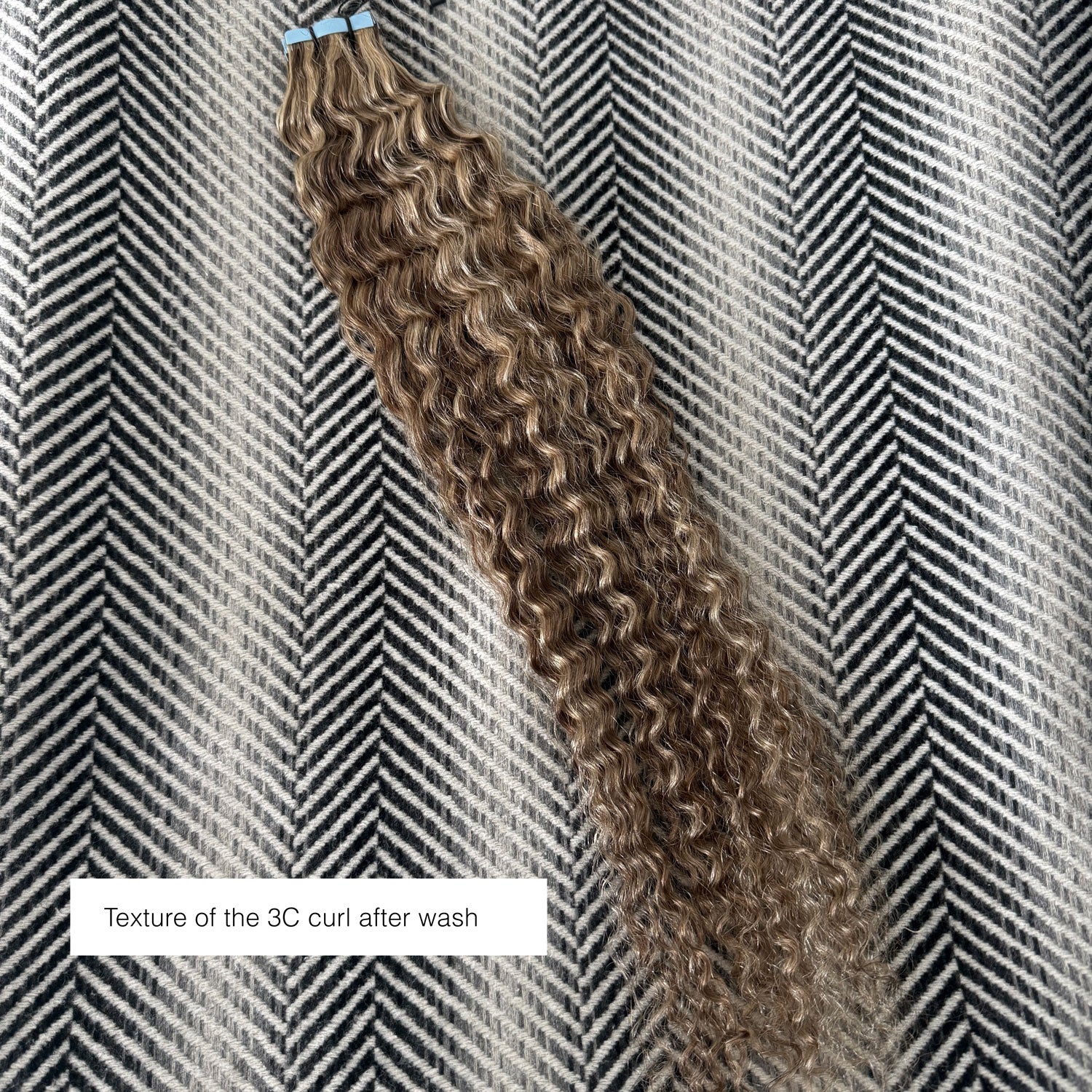 A bundle of 3C curly hair extensions in a medium brown color, displayed diagonally on a black and white herringbone patterned fabric. A text overlay at the bottom left reads 'Texture of the 3C curl After Wash'.