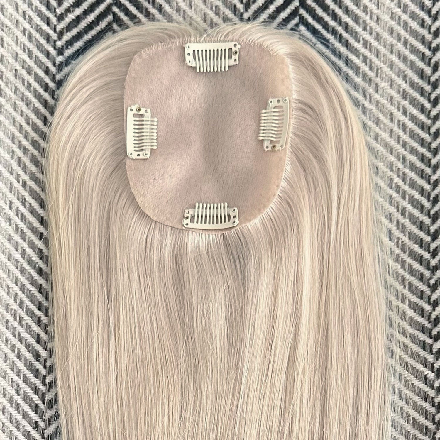 A light ash blonde clip-in hair extension piece with four silver metal clips, laid flat on a gray and white herringbone patterned fabric background.