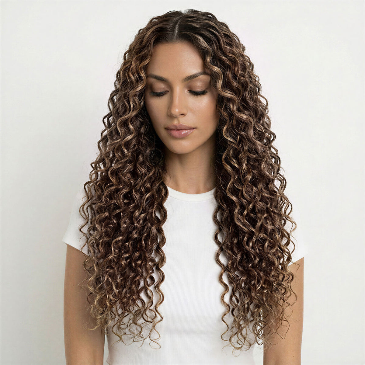 Woman with long, wavy brown and caramel curly hair extensions wearing a white shirt on a light background