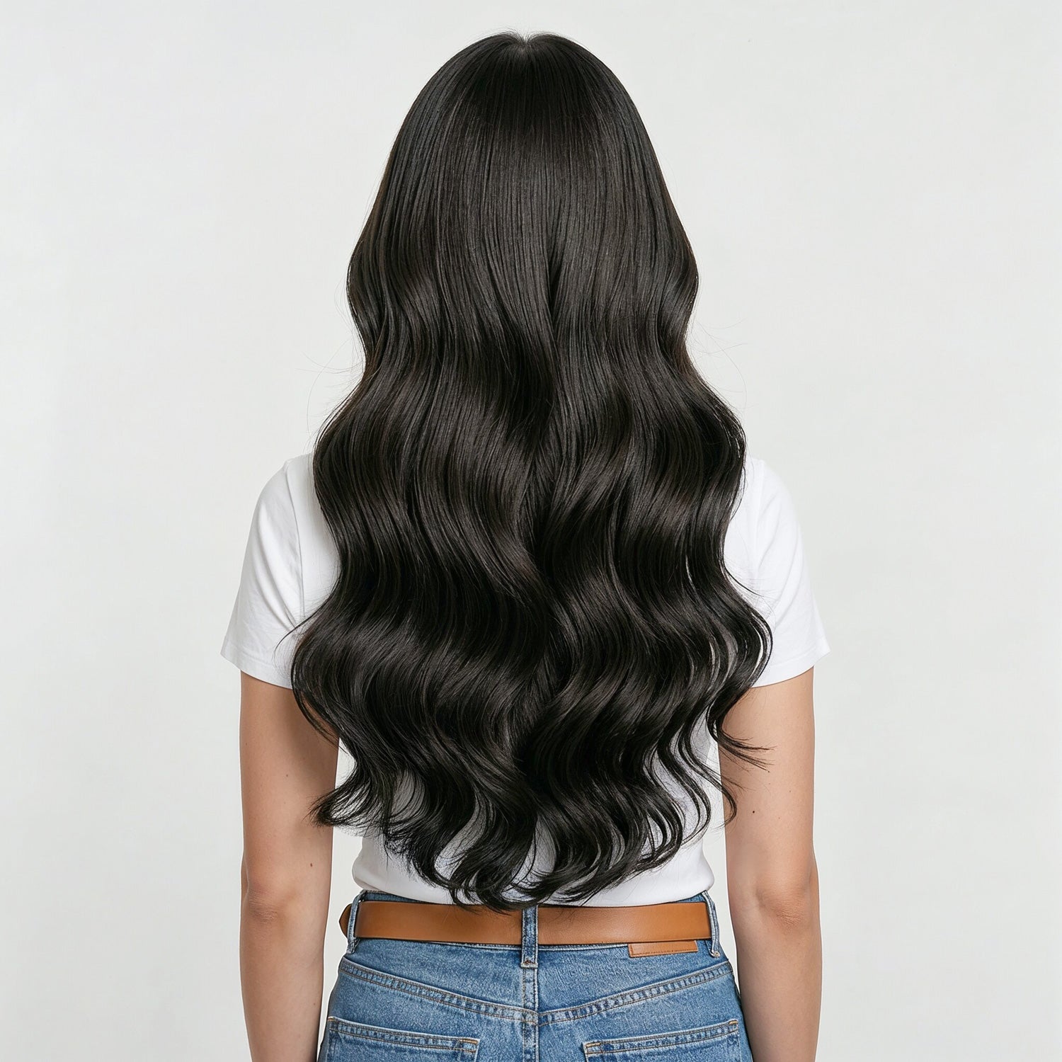 Person with long, wavy black hair extensions wearing a white shirt and blue jeans on a light gray background