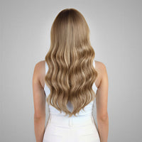 A woman viewed from behind wearing a white sleeveless top and white pants, showcasing long, wavy blonde hair against a neutral gray background.