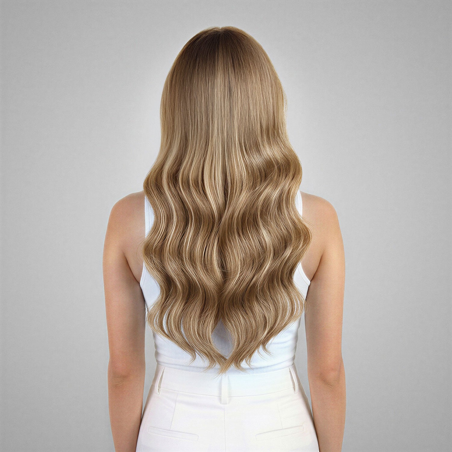 A woman viewed from behind wearing a white sleeveless top and white pants, showcasing long, wavy blonde hair against a neutral gray background.